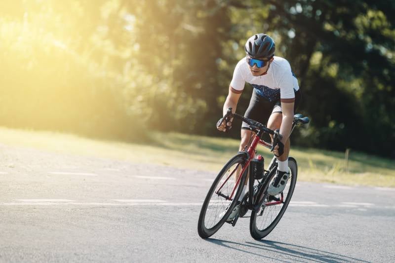Cyclist in a helmet and sunglasses riding a red road bike on a sunny paved road.