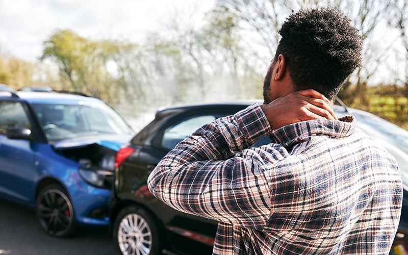 Man holding his neck in pain after a car accident, with two damaged cars visible in the background.