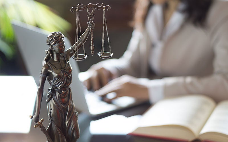 Lady Justice statue in front of a blurred person typing on a laptop with an open book.