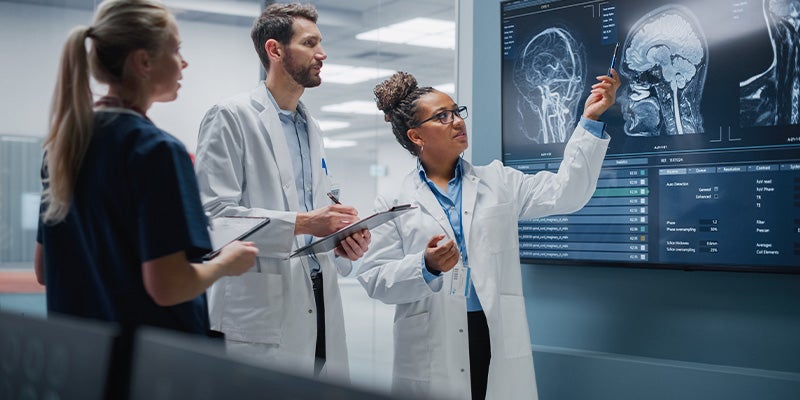 Three medical professionals examine brain scans on a large digital display, with one pointing at the screen.