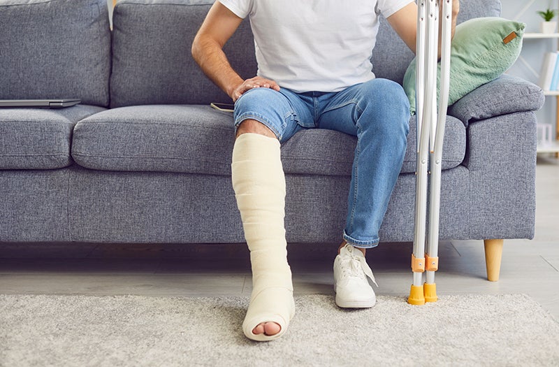 Person sitting on a couch with one leg bandaged or in a cast, and crutches beside them.