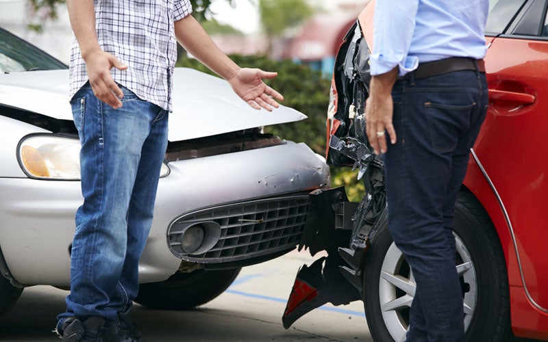 Two men stand next to damaged cars after a rear-end collision. One man gestures with his hands in frustration.