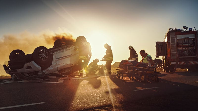Emergency responders at a fiery car crash with an overturned car and a person on a stretcher at sunset.