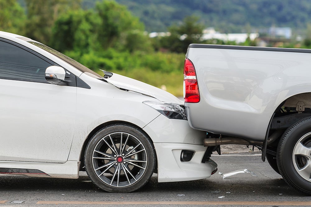 White car with crumpled hood and front bumper after rear-ending a silver pickup truck.