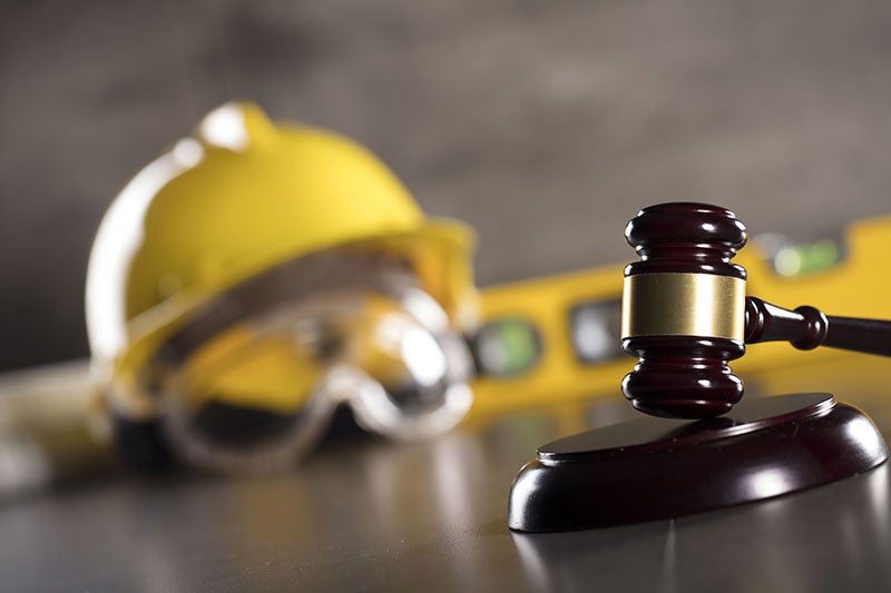 Gavel in foreground, with a blurry yellow hard hat, safety goggles, and construction level in the background.