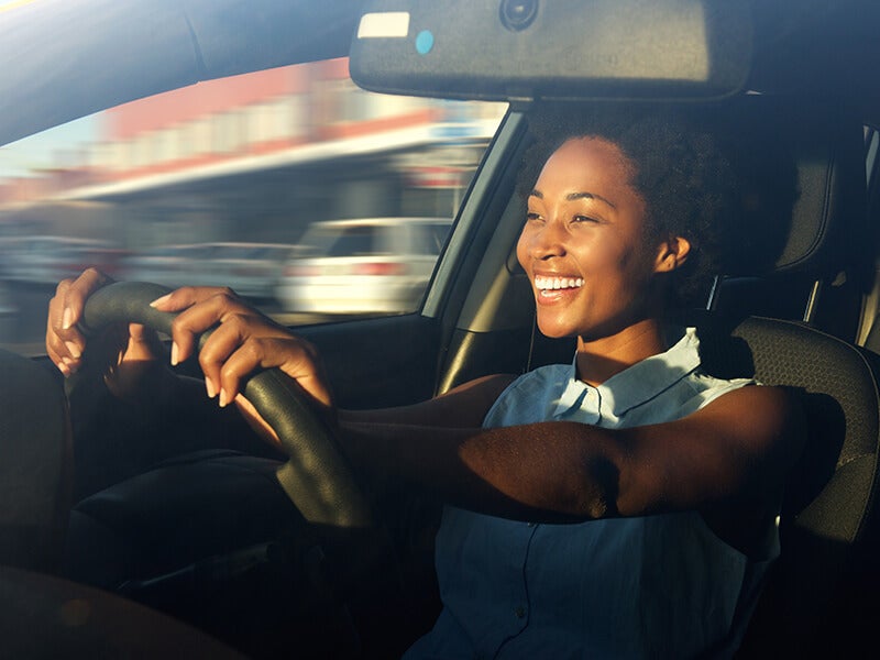 Smiling Black woman driving a car, hands on the wheel, sunlight on her face, with motion blur in the background.