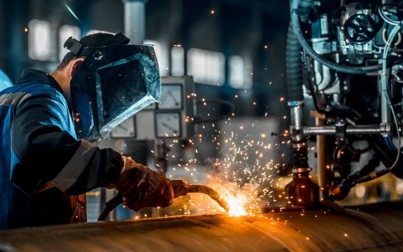 Welder in a helmet and gloves creating bright sparks while working on a large metal pipe.