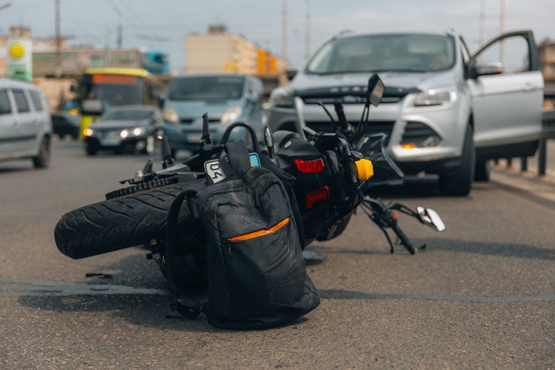 A fallen black motorcycle and backpack on a road after an accident, with a silver SUV and other vehicles in the background.