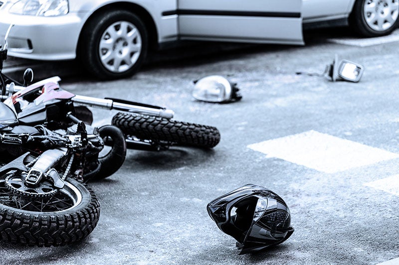 A motorcycle lies on the ground next to a car with an open door, with a helmet and car debris scattered on the road.
