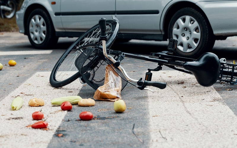 A bicycle and groceries lie spilled on a crosswalk, with a silver car visible in the background, suggesting an accident.