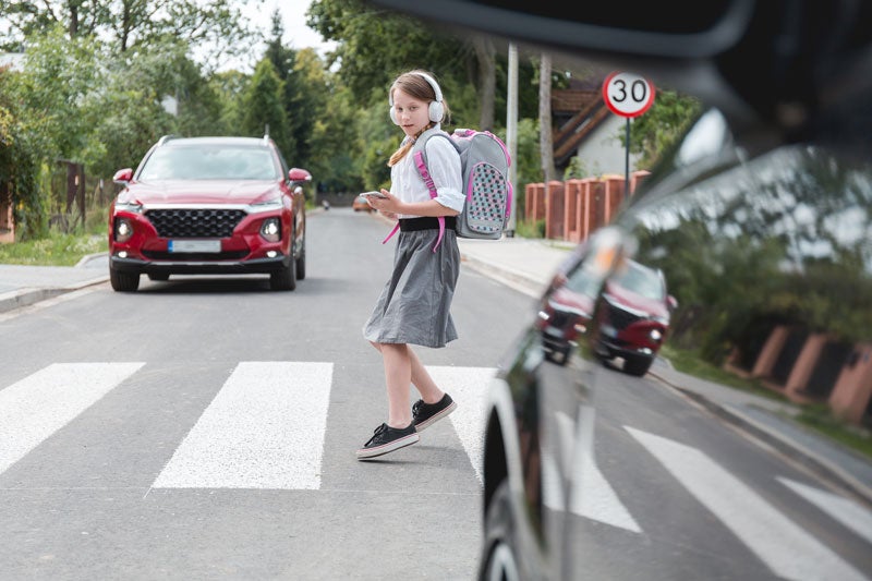 Girl with headphones and backpack crossing street while looking at phone, red car stopped at crosswalk.