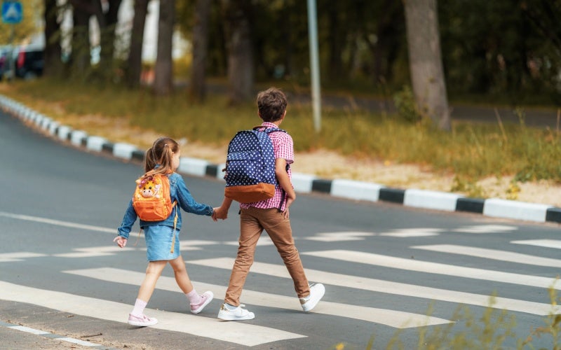 Two children with backpacks, holding hands, walk across a zebra crossing.
