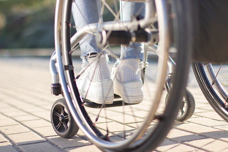 Person in a wheelchair, seen through the wheels, wearing white sneakers and ripped jeans on a paved surface.