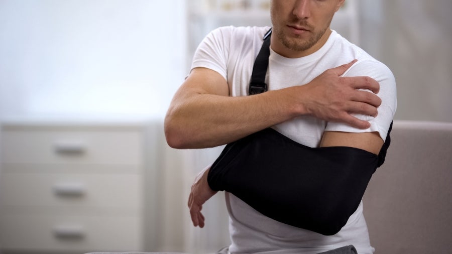 Man with a black arm sling holding his injured shoulder, looking uncomfortable.