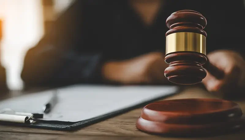 A wooden gavel hovers above its sounding block on a desk, with a clipboard to the left and a person in the background.