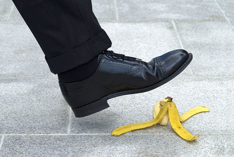 A person's foot in a black dress shoe is about to step on a banana peel on a grey sidewalk.