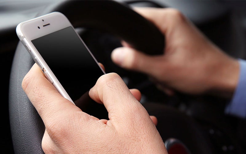 Person holding a smartphone with one hand and a steering wheel with the other.