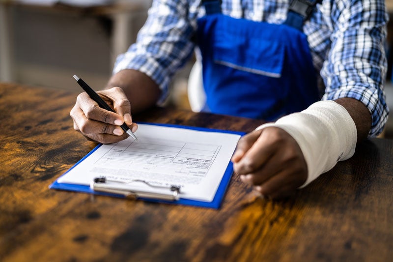 Worker in blue overalls with a bandaged left wrist, writing on a document on a clipboard.