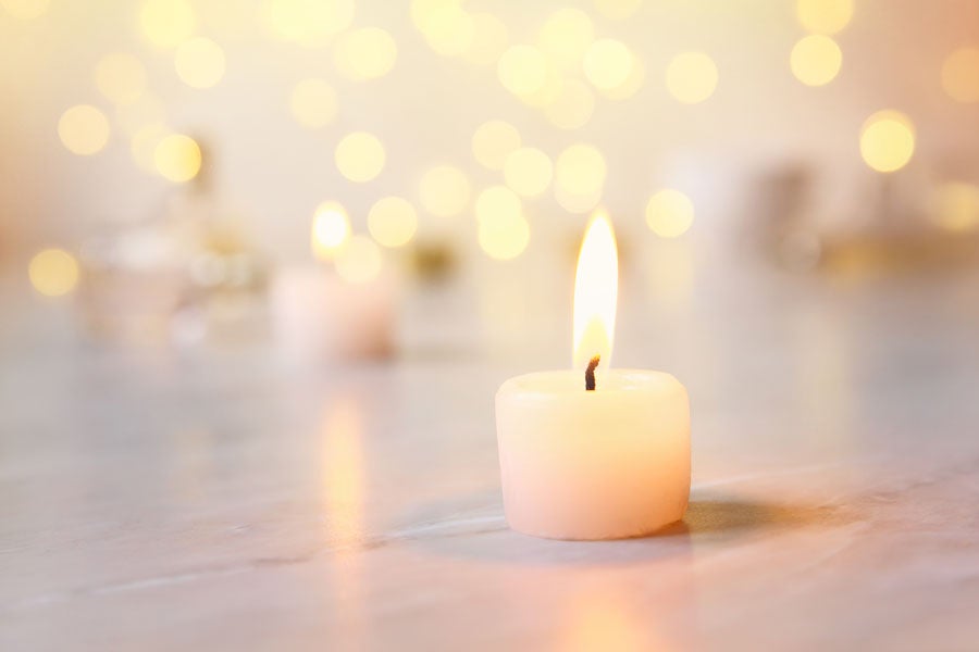 A single small, lit candle on a light surface with blurred warm bokeh lights in the background.